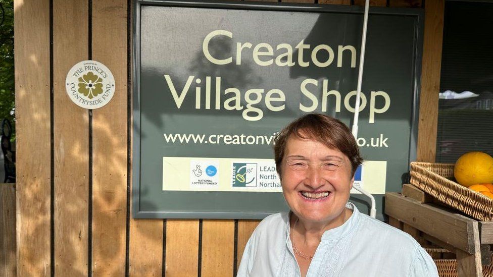 Smiling woman wearing light top stands outside village shop