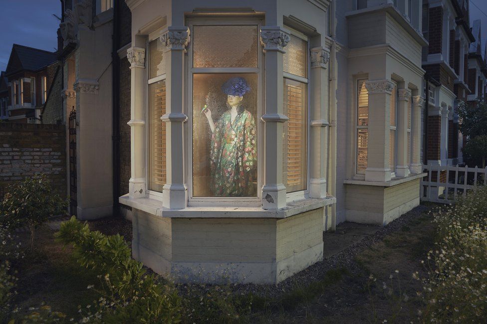 A woman stands looking out the window of a house in London in a stylised portrait