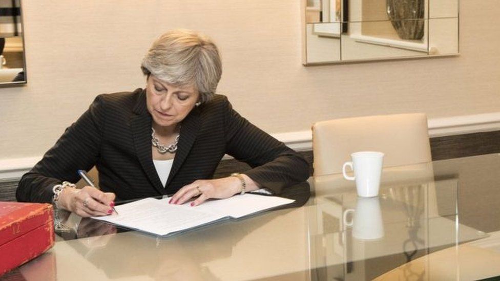 Theresa May sitting at desk singing papers