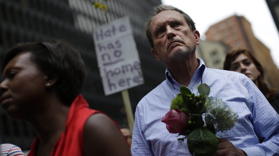 a man holding flowers in front of a sign that reads "Hate has no home here"