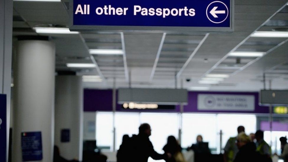 Airport sign reading 'all other passports' with a directional arrow, with passengers in the background