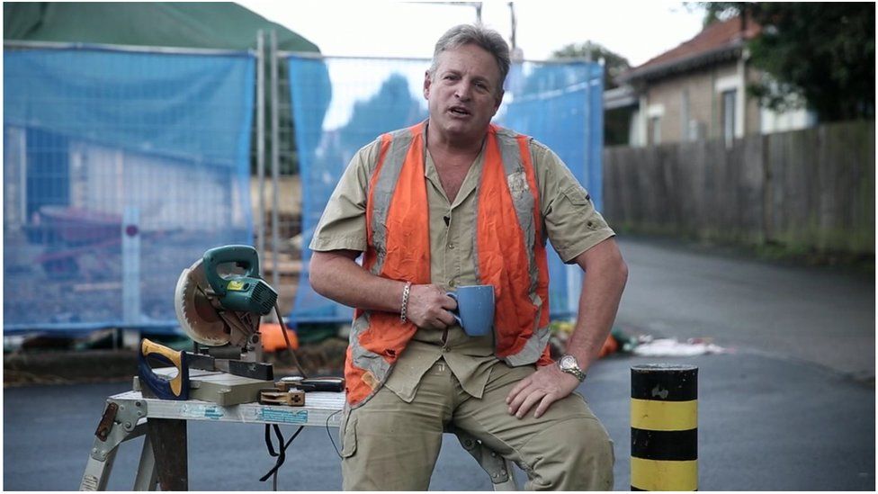 Man dressed as tradesman speaks in still from an advertisement