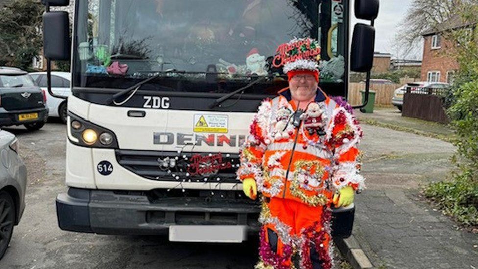 Man with big Christmas crown and red costume covered with lights and other decorations stands in front of a bin lorry