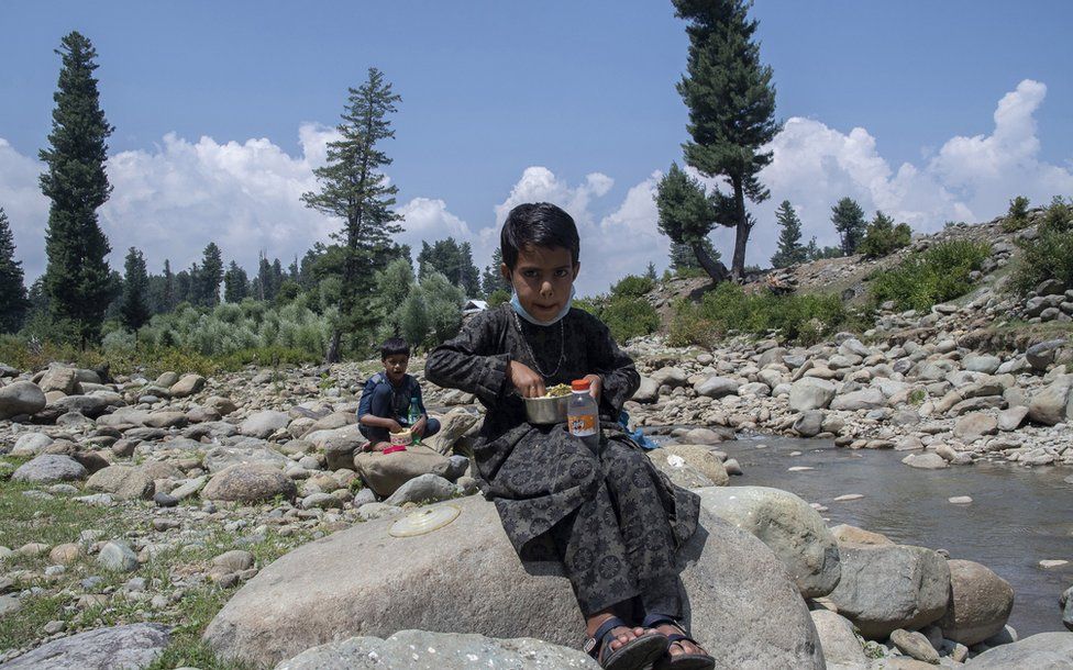 A boy eats lunch sitting on a rock by a stream.
