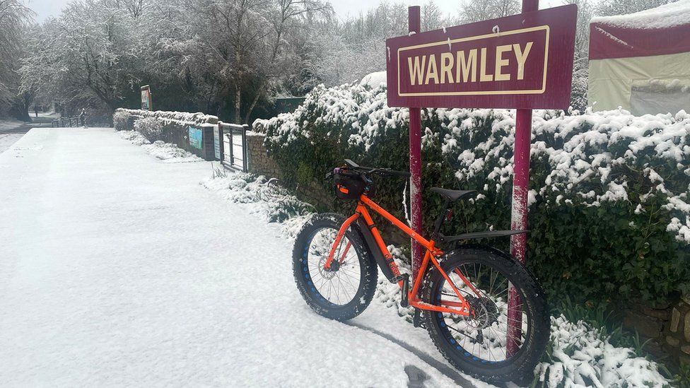 A mountain bike parked next to a sign saying Warmley, with snow on the ground and hedgerow