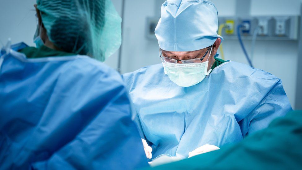 A surgeon wearing glasses and a face mask is seen looking down while performing an operation