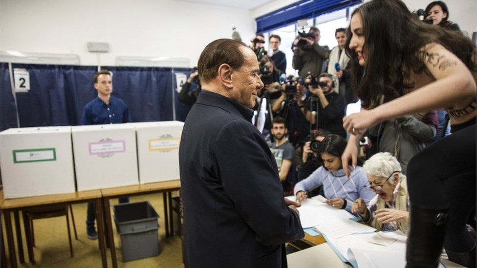Mr Berlusconi stands in front of press as a woman standing on a table, with slogans written on her topless torso