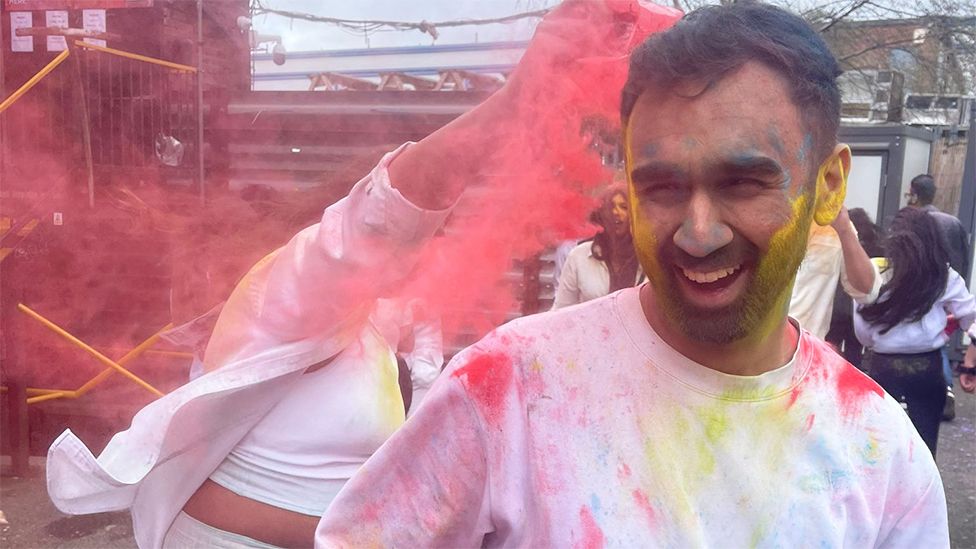 Almir, a man, wearing a white tshirt which has red and yellow powdered colour on it. He is smiling, with his beard coloured yellow and someone behind him throwing red powdered colour on him as they celebrate.
