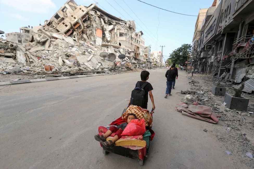 A Palestinian boy pulls a cart carrying his brother and their belongings as they flee their home during Israeli air and artillery strikes, near the site of destroyed a tower block in Gaza City (14 May 2021)