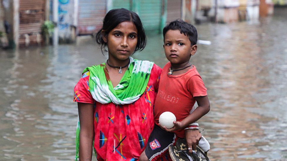 A woman holding her child, stands in a waterlogged street due to the high tide in the river Ganges.