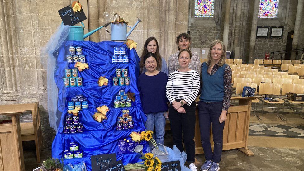 Five people stood next to the water is life display, which features blue fabric draped over a stair-like structure with tinned food and flowers on it