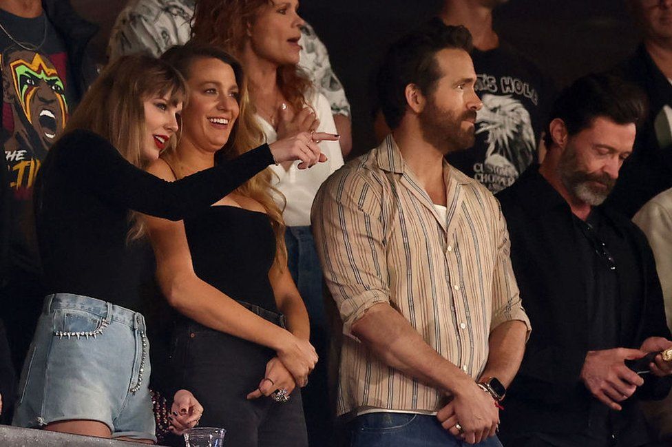 Taylor Swift and Blake Lively smile as the singer points towards something, with Ryan Reynolds and Hugh Jackman in the stand just to their right