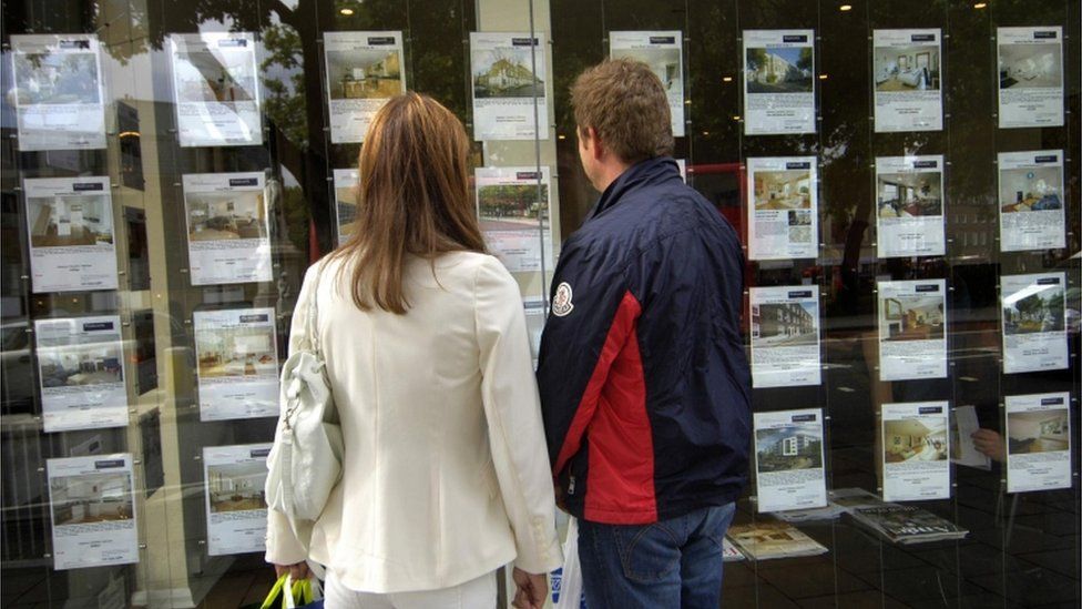 A couple, a woman in a white jacket and a man wearing a windbreaker, standing outside an estate agent looking in the window