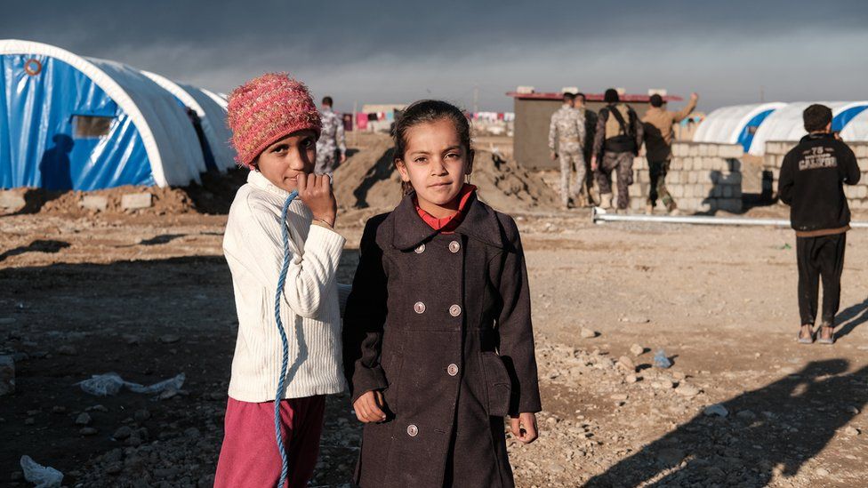 Two girls look at the camera amid a refugee camp with the blue tents of UNICEF visible nearby