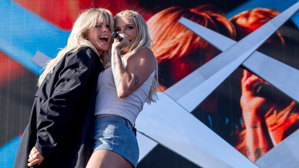 Reneé Rapp and Kesha performing on stage at Coachella. The pair are standing incredibly close together, with Reneé grinning and Kesha singing
