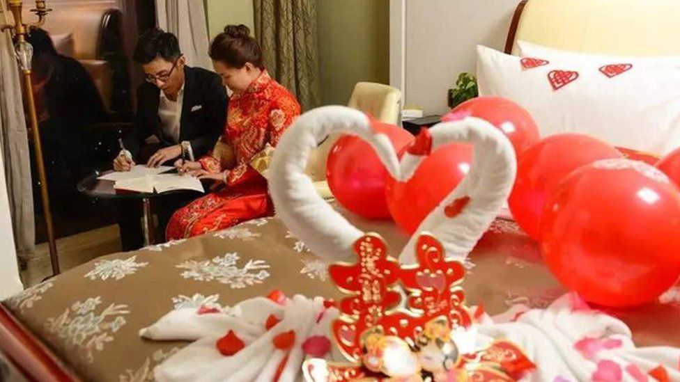 A Chinese couple sits on a desk beside their matrimonial bed