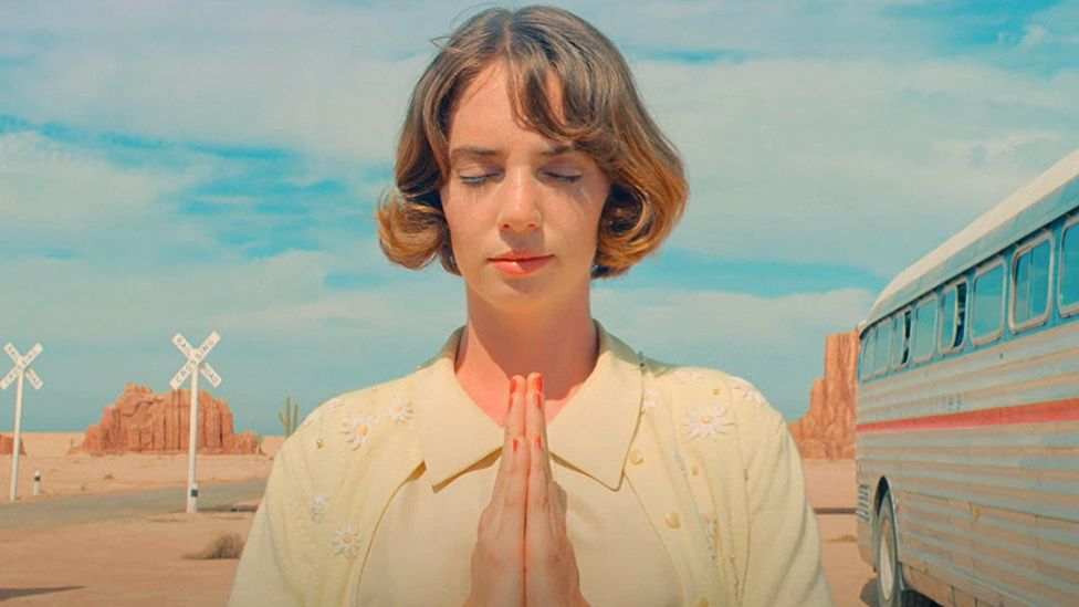 Maya Hawke clasps her hands with her eyes closed with open sky and road background in Asteroid City