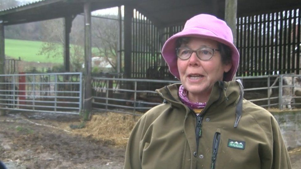 Susan Mannerings wearing a pink hat and fleece standing in front of a cowshed