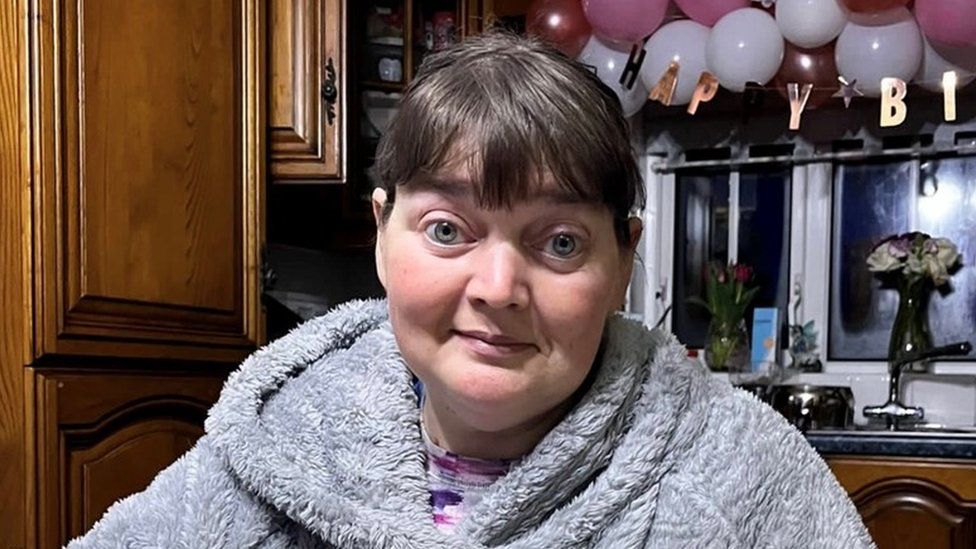 Pamela Bloomer, wearing a grey dressing gown, stands in her kitchen