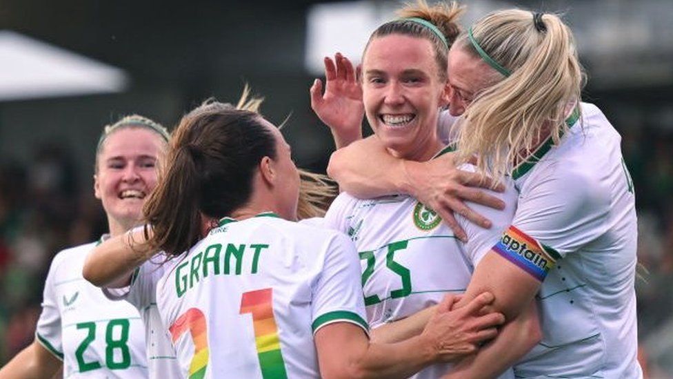 Claire O'Riordan celebrating with her Republic of Ireland team-mates, from left to right Erin McLaughlin, Ciara Grant and Louise Quinn after scoring a goal. They are all smiling and wearing their Ireland kit which is white shirts and green shorts