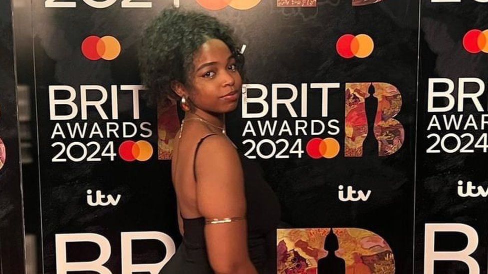 Nayana Brathwaite pictured on the Brits red carpet. Nayana is a 21-year-old black woman who wears her hair styled in an up do. She stands facing to the right, looking at the camera over her shoulder. She wears a strappy black dress and large gold earrings and arm bangle.