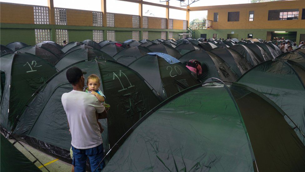 A man holds his baby while looking down a row of tents set up in a shelter