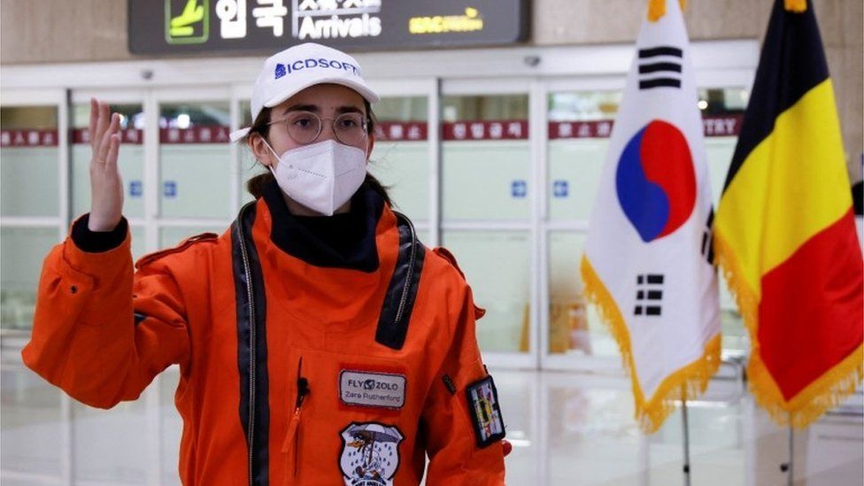 Zara Rutherford standing in arrivals at Gimpo International Airport in Seoul wearing an orange immersion suit and a white facemask while she is interviewed by journalists