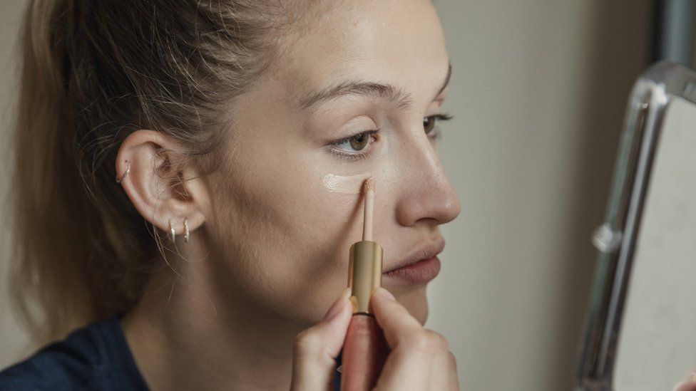 Woman applying concealer under eyes in front of hand held mirror