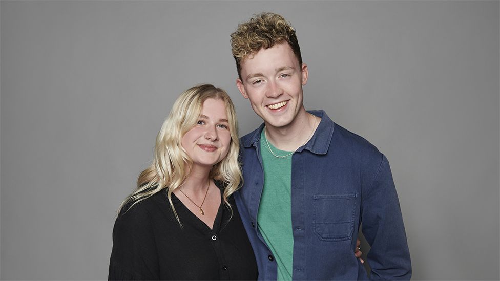 Danni Diston is on the left and Sam MacGregor is on the right. They are standing next to each other, smiling looking at the camera with a plain grey background. Danni is wearing a black top, while Sam has a green top with a dark blue jacket over the top.
