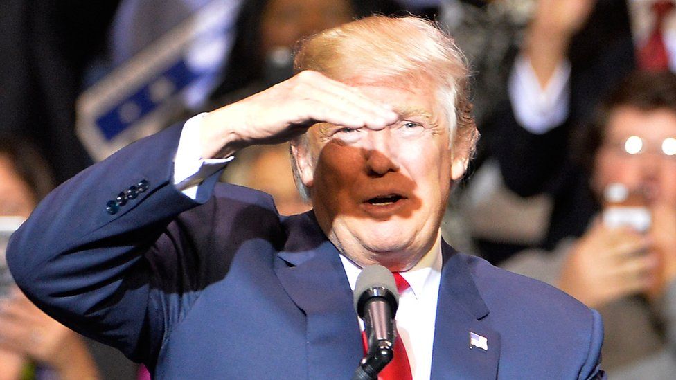 Donald Trump raises his hand to shield his eyes from a bright ray of light