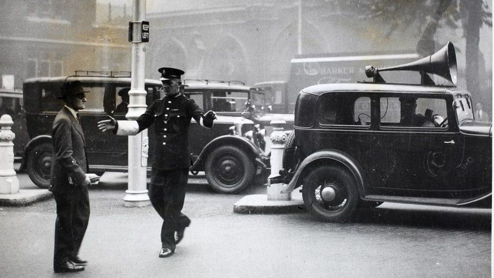An erring pedestrian at Kensington Church Street is severely lectured by a traffic policeman for not using a crossing correctly in the mist, London, England. The morning's mist in London gave the police authorities an opportunity of testing out a new loudspeaker as an additional method of traffic and pedestrian control at busy points. The loudspeaker is attached to a van manned by driver and microphone attendants, who can issue instructions to both traffic and pedestrians as required, if, in the words of a police official, 'they need instruction'. In some cases the driver of a vehicle will be stopped and given a lecture on road manners and public safety. Pedestrians are also liable to these lectures. (Photo by Fox Photos/Hulton Archive/Getty Images)