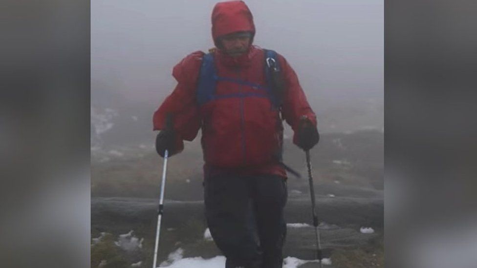 Ian Evans participating in the Winter Spine Challenge in January 2023. He is wearing a red coat and walking with hiking sticks. He is along the Pennine Way and a small amount of snow can be seen on the ground.