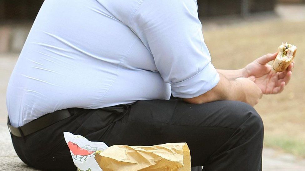 An obese man eating a sandwich while sat outside