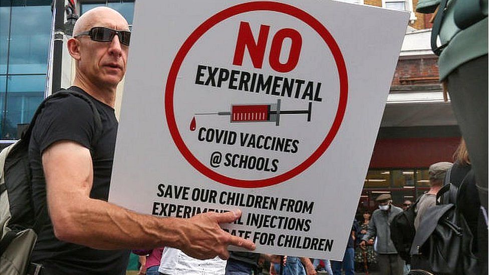 man holding sign reading 'no experimental covid vaccines @schools'