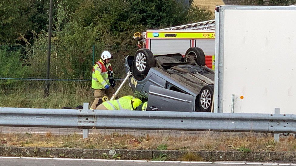 Men in high vis attending to an upturned car with a fire truck behind it