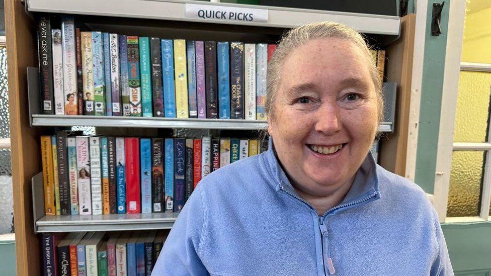Smiling woman in blue sweater stands in front of book shelves