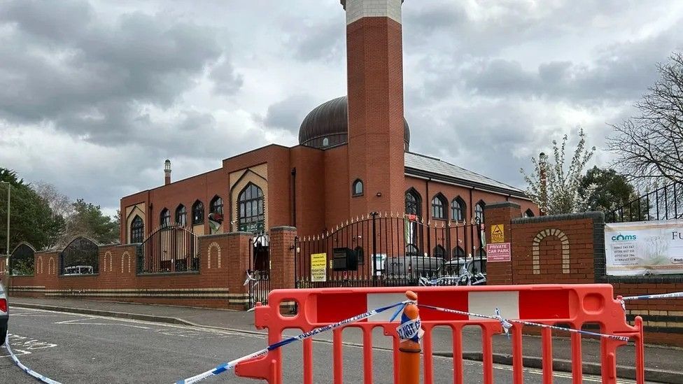 Manzil Way, with a cordon of police tape, and barriers, in the foreground. The mosque, made of brown brick, is on the other side of the road.