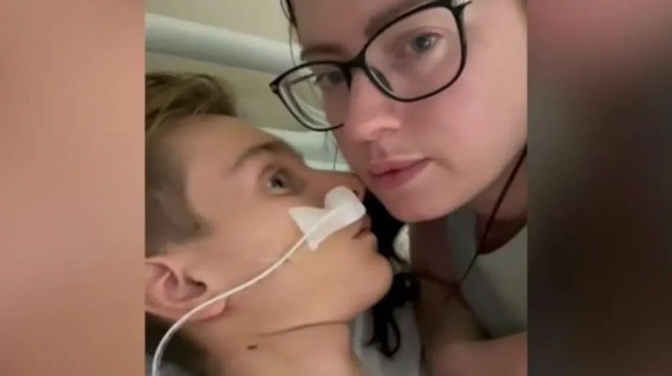 A still from a video showing Jack Dolan with his face close to his mother in hospital. She is looking at the camera.