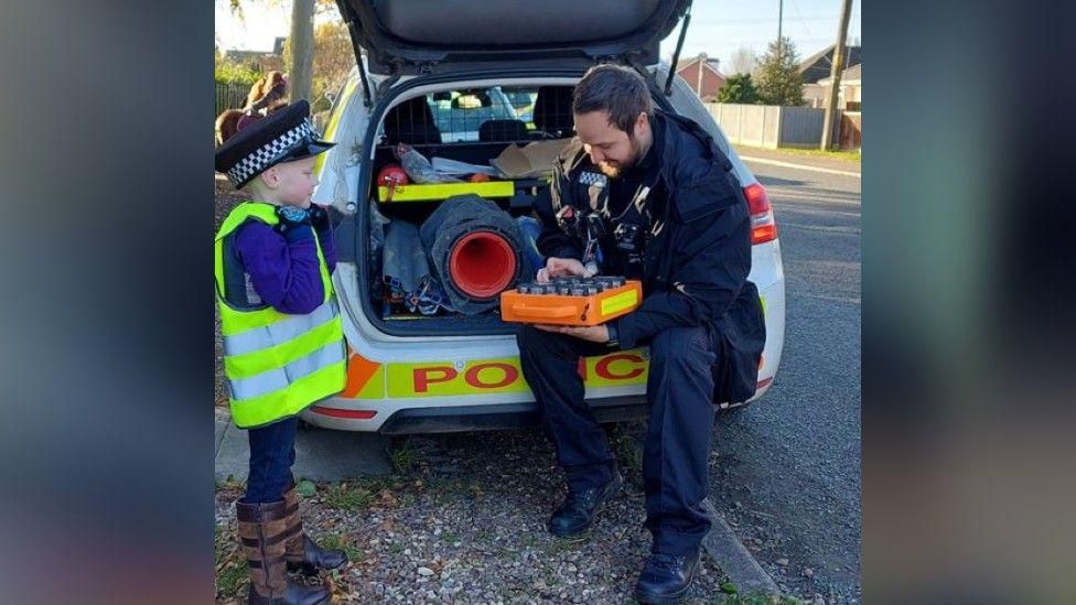 A policeman showing Kodie how their police equipment works in the boot of the car. 