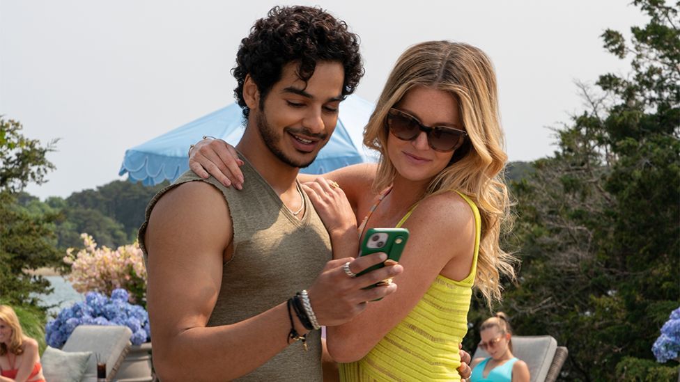 A still from Netflix show The Perfect Couple with Ishaan's character Shooter wearing a light brown or green vest and holding a phone as the character of Meritt, wearing a yellow top, puts her arm around him. There are green trees and landscape in the background.