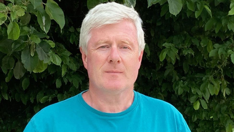 George Byrne wearing a turquoise t-shirt and looking against a dark green leafy hedge
