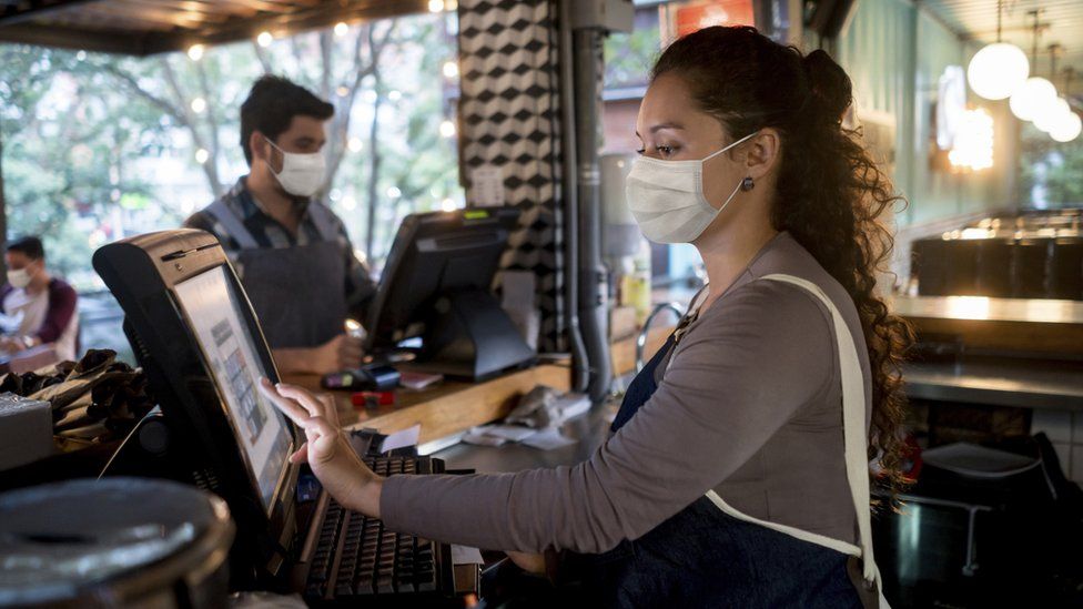 A woman working at the cashier at a restaurant wearing a facemask