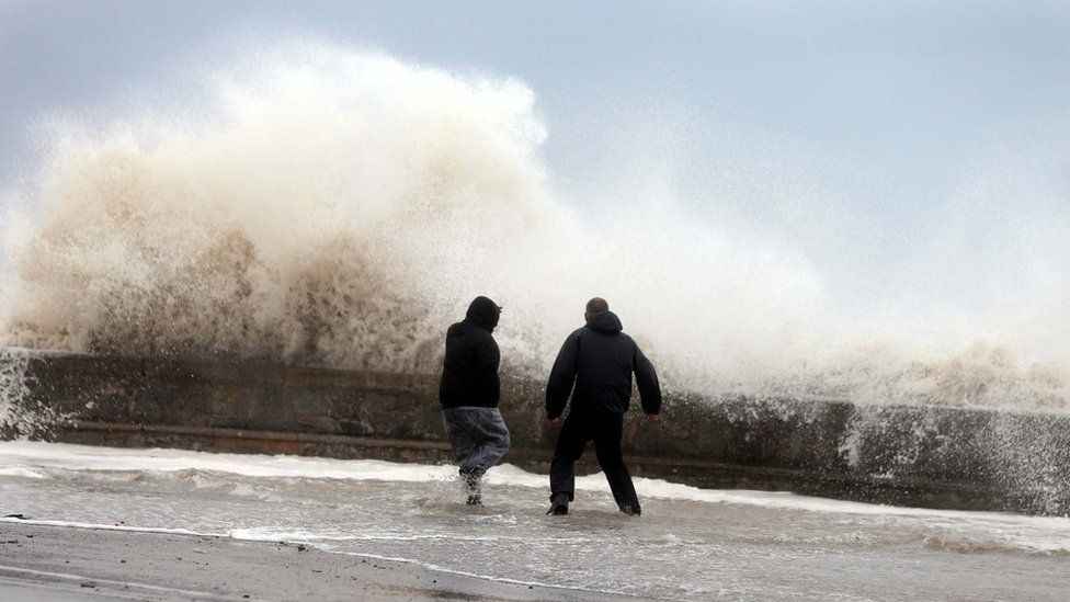 Two men standing on a sea wall as a wave breaks