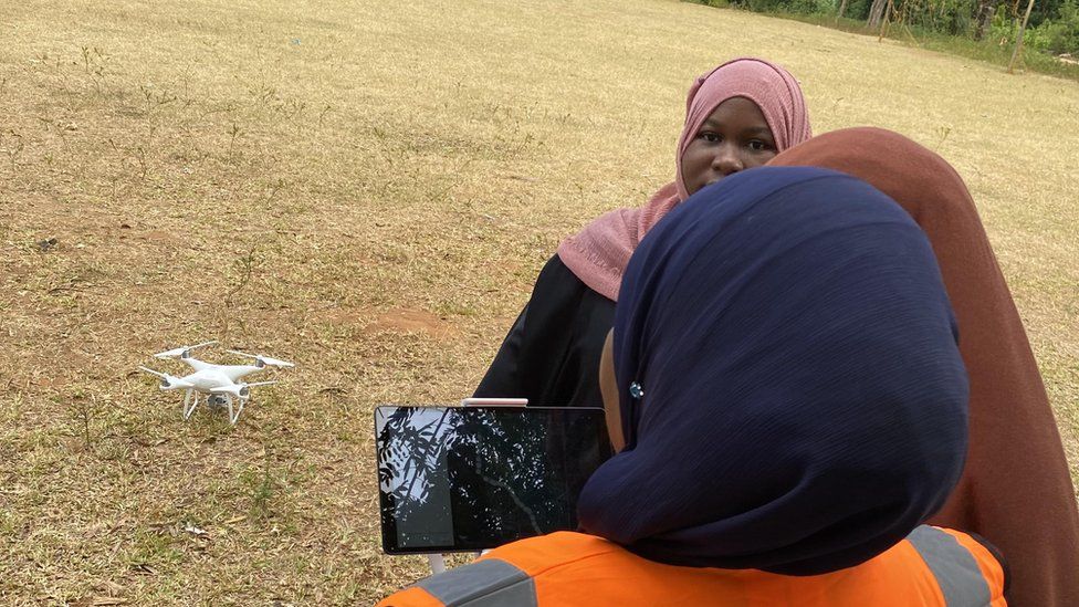 Three Zanzibar women in head scarves hold a smartphone with a drone on the ground beyond them