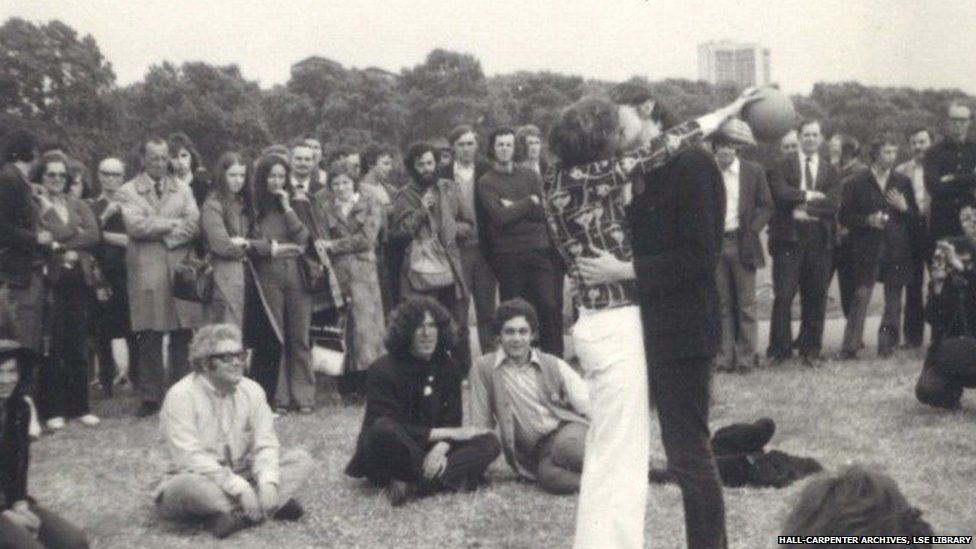 Black and white photograph of two men kissing in a park while a group watch on