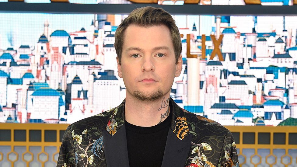 A man with neatly cropped hair stands in front of a large picture of a town made up of tall, bright white buildings. It looks Meditteranean. He has handwriting tattooed on his neck and wears a black blazer with a traditional Japanese pattern featuring cranes and paper fans.