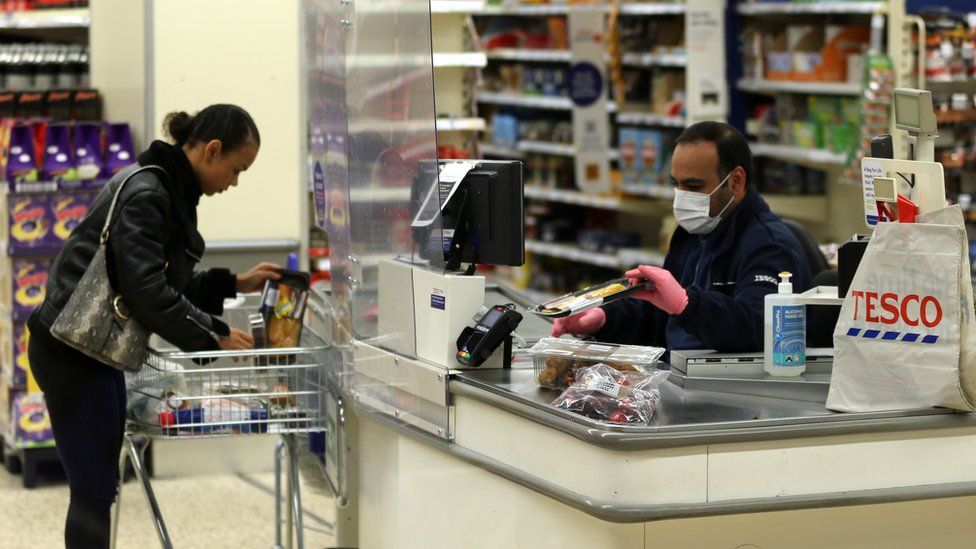 A Tesco supermarket cashier wearing protective face mask and gloves assists a shopper behind a plastic screen