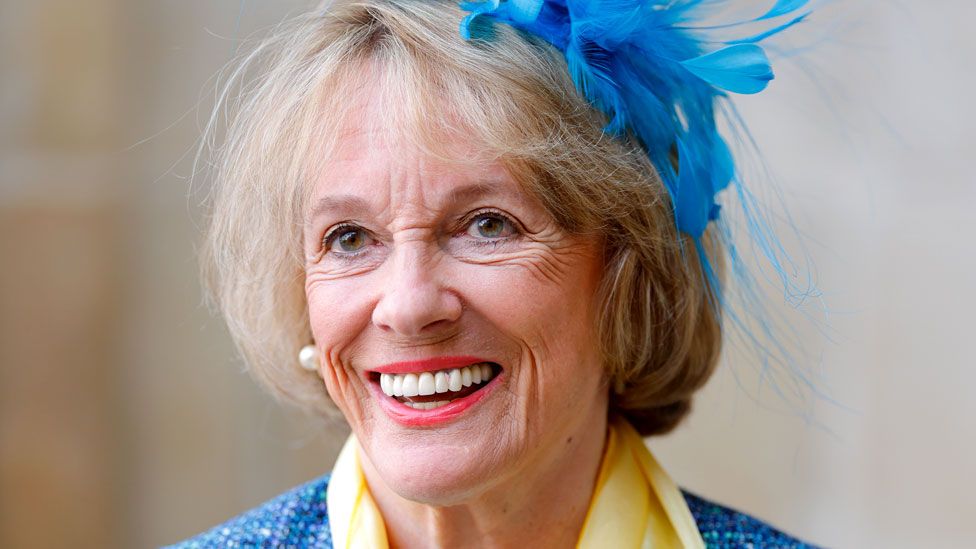 Dame Esther smiles, dressed up with a feathered blue fascinator