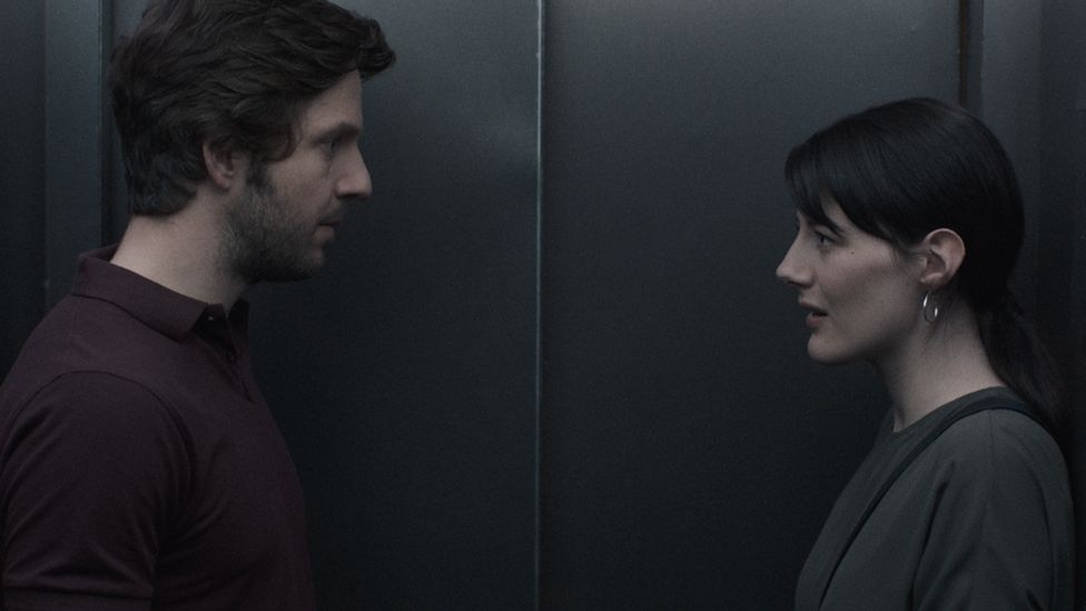 A still from a film. A man and woman stare at one another inside a lift.