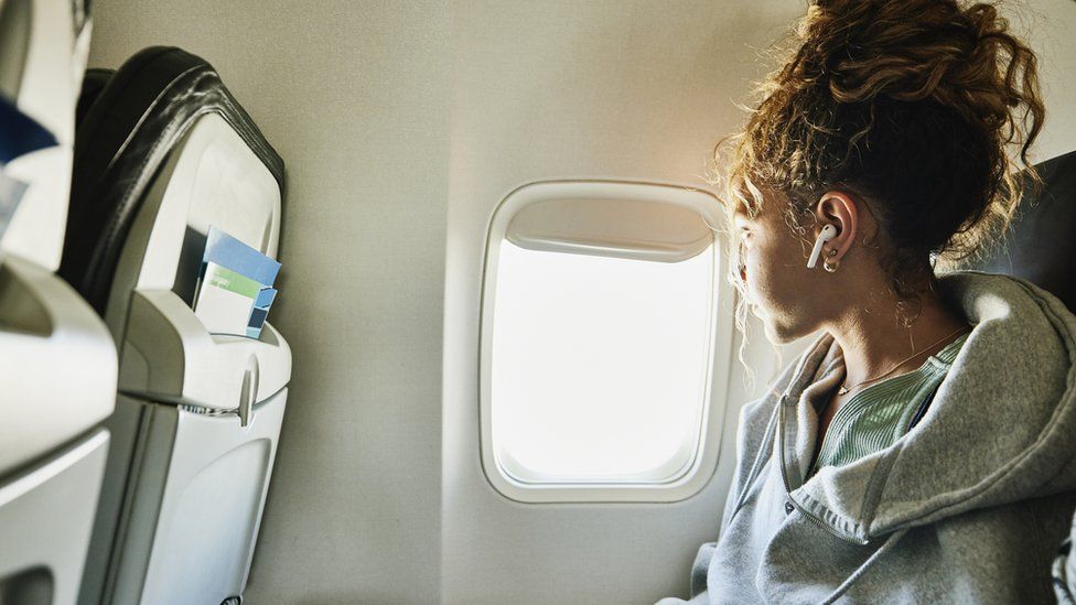 Generic image of a young woman on a commercial flight. She wears a grey hoodie and has curly brown hair tied back in a messy bun. She has headphones in her ears and is looking out of the window. A row of seats is visible in front of her
